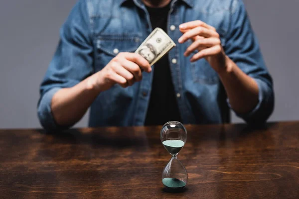 Foco selectivo del reloj de arena cerca del hombre sosteniendo dólares mientras está sentado en la mesa aislado en gris, concepto de gestión del tiempo - foto de stock