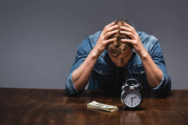 Homem insatisfeito olhando para o dinheiro e despertador na mesa isolado em cinza, conceito de gerenciamento de tempo — Fotografia de Stock