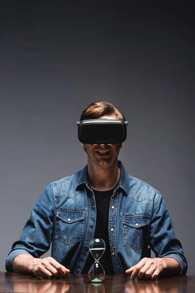 Selektiver Fokus eines lächelnden Mannes im vr-Headset, der neben Sanduhr auf Tisch auf grauem Hintergrund sitzt, Konzept des Zeitmanagements — Stockfoto