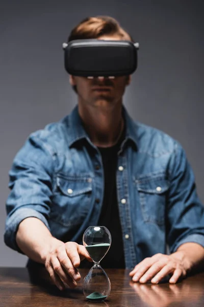 Enfoque selectivo del hombre en los auriculares de realidad virtual tirando de la mano al reloj de arena en la mesa sobre fondo gris, concepto de gestión del tiempo - foto de stock