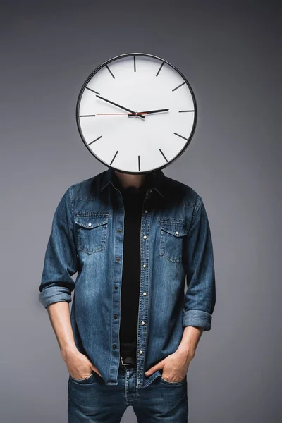 Young man with clock on head and hands in pockets of jeans on grey background, concept of time management — Stock Photo