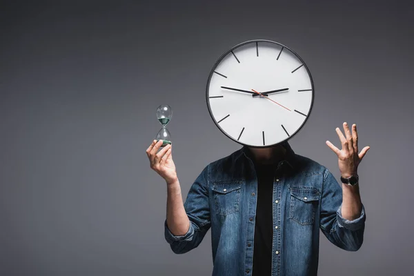 Hombre con reloj en la cabeza sosteniendo reloj de arena y gestos sobre fondo gris, concepto de gestión del tiempo - foto de stock