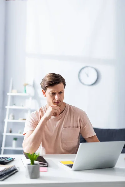 Selective focus of pensive freelancer looking at laptop near stationery on table in living room, earning online concept — Stock Photo