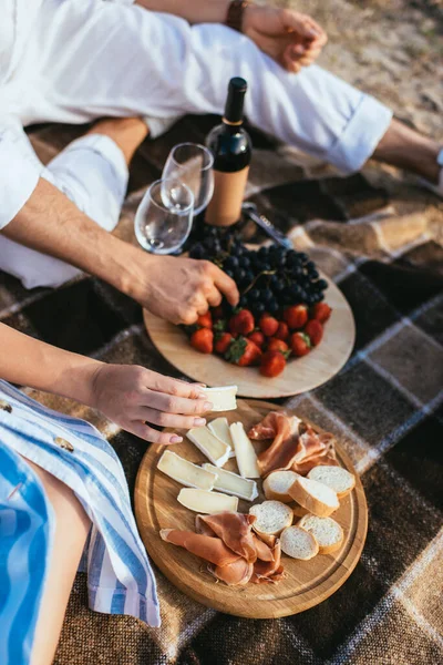 Cropped view of couple near fruits, soft cheese and glasses on plaid blanket — Stock Photo