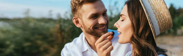 Tiro panorámico de hombre feliz sosteniendo uva cerca de chica con los ojos cerrados - foto de stock