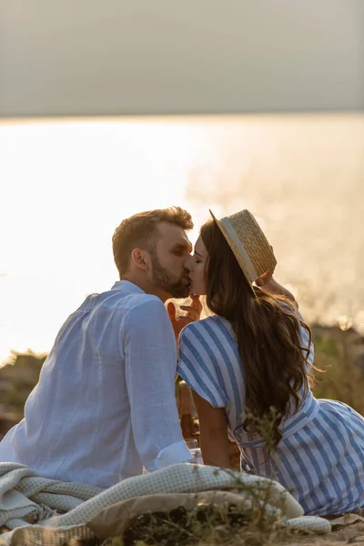 Vista lateral de hombre guapo y mujer atractiva en sombrero de paja besándose cerca del lago - foto de stock