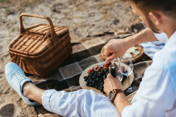 Selektiver Fokus des bärtigen Mannes, der eine Flasche Wein in der Nähe von Früchten auf dem Teller und einen Weidenkorb auf der Decke öffnet — Stockfoto