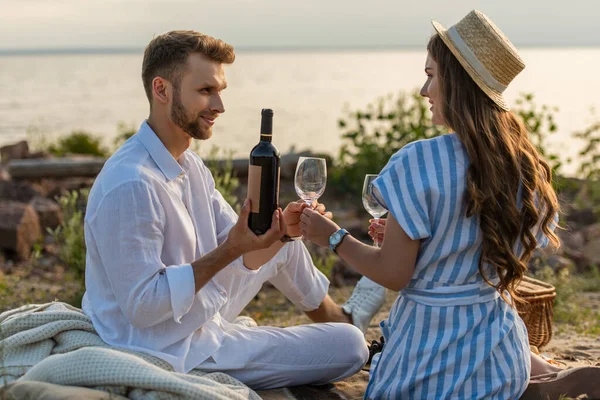 Barbudo hombre celebración botella de vino y mirando feliz mujer en paja sombrero sentado en manta - foto de stock