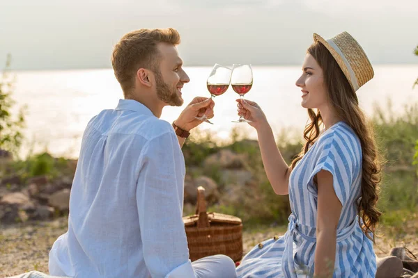 Vista lateral de alegre pareja tintineo vasos con vino tinto mientras está sentado cerca del lago - foto de stock