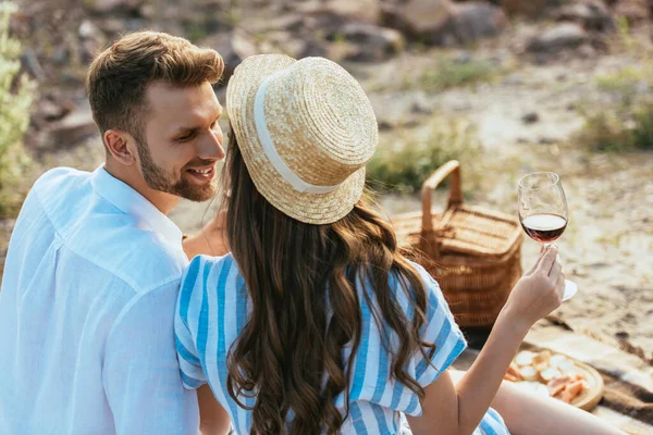 Selektiver Fokus des Mädchens mit Strohhut, das Glas mit Rotwein in der Nähe des glücklichen Freundes hält — Stockfoto