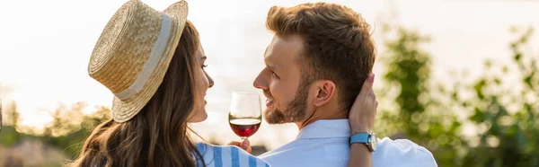 Récolte panoramique de femme gaie en chapeau de paille touchant petit ami heureux près du verre avec du vin rouge — Photo de stock