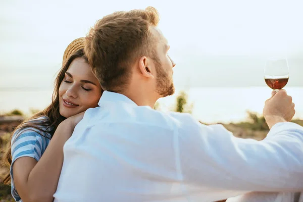 Cheerful woman in straw hat hugging boyfriend holding glass with red wine — Stock Photo