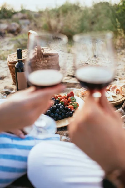 Selective focus of plate with fresh fruits near bottle and couple clinking glasses with red wine — Stock Photo