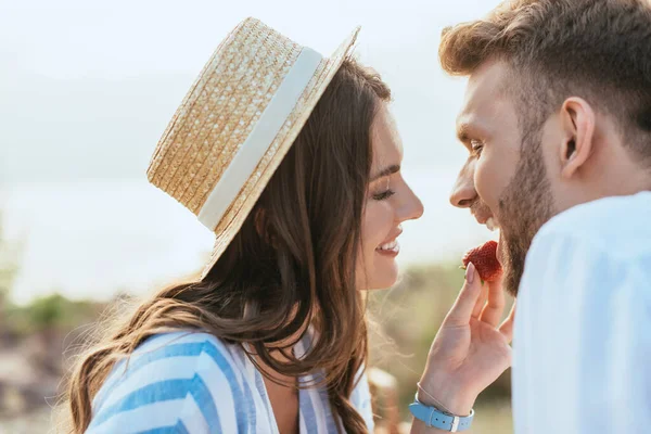 Foco seletivo da mulher feliz em chapéu de palha alimentando namorado com morango maduro — Fotografia de Stock