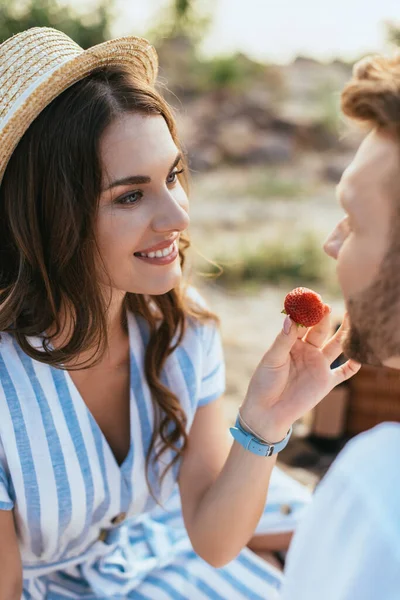 Foco seletivo da mulher feliz em chapéu de palha namorado de alimentação com morango — Fotografia de Stock