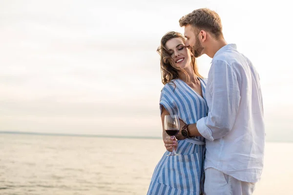 Happy girl holding glass with red wine near cheerful boyfriend while standing near lake — Stock Photo