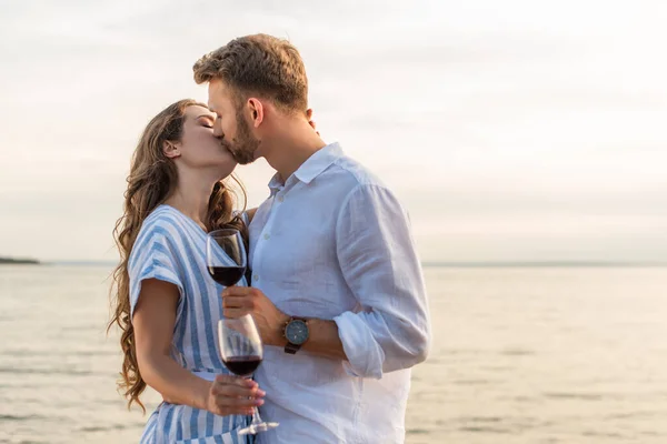 Casal beijando e segurando copos com vinho tinto perto do lago — Fotografia de Stock