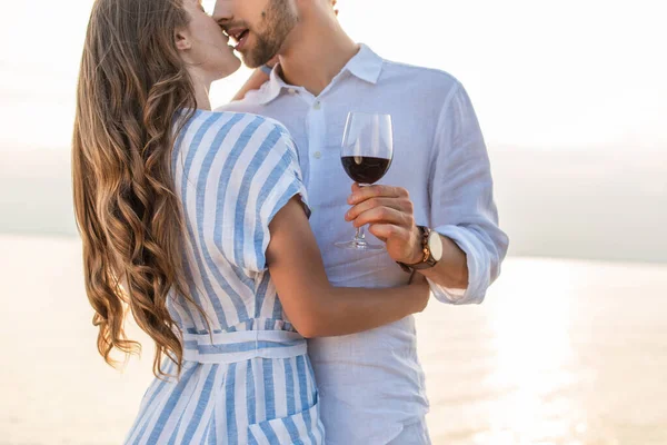 Cropped view of bearded man kissing girlfriend and holding glass with red wine near lake — Stock Photo