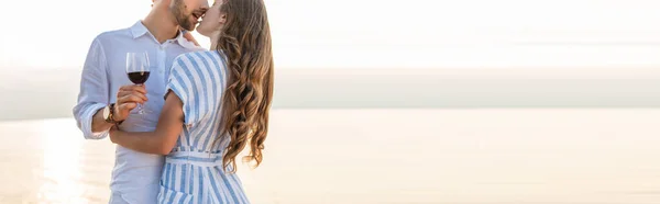 Horizontal crop of bearded man kissing girlfriend and holding glass with red wine near lake — Stock Photo