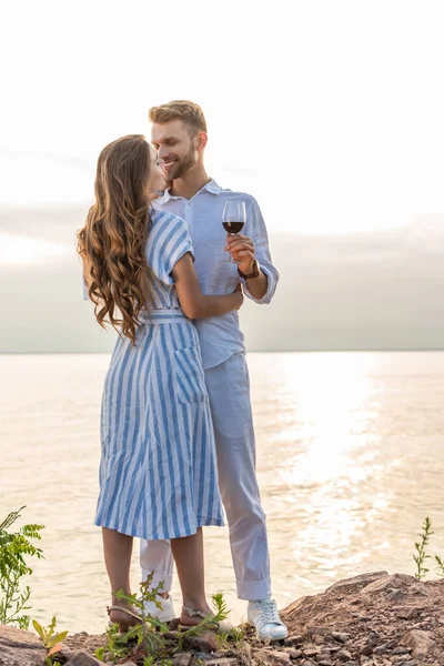 Happy man holding glass with red wine near cheerful girl while standing near lake — Stock Photo