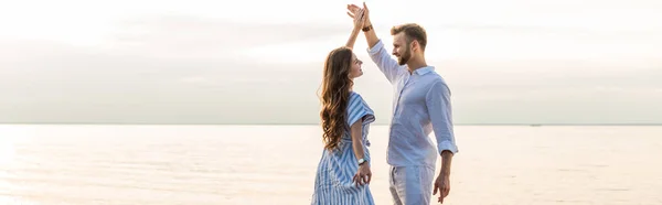Панорамный снимок счастливой пары, держащейся за руки и танцующей у озера — стоковое фото