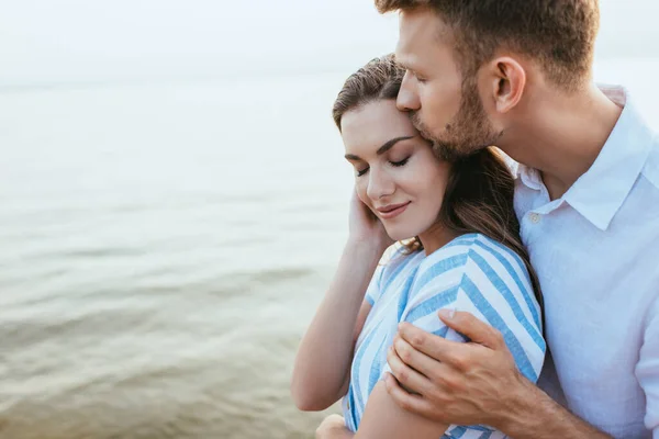 Бородатый мужчина целует красивую девушку, улыбающуюся у реки — стоковое фото
