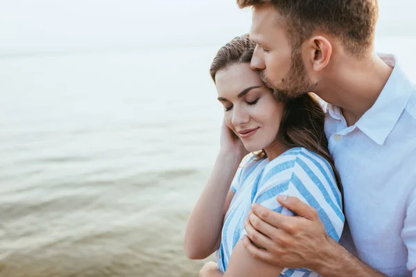 Barbudo hombre besos hermosa novia sonriendo cerca de river - foto de stock