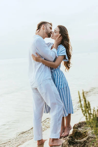 Selective focus of happy man hugging and kissing girl in dress near lake — Stock Photo