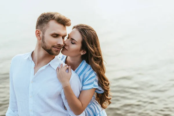 Mujer atractiva abrazando hombre guapo con los ojos cerrados cerca del río - foto de stock