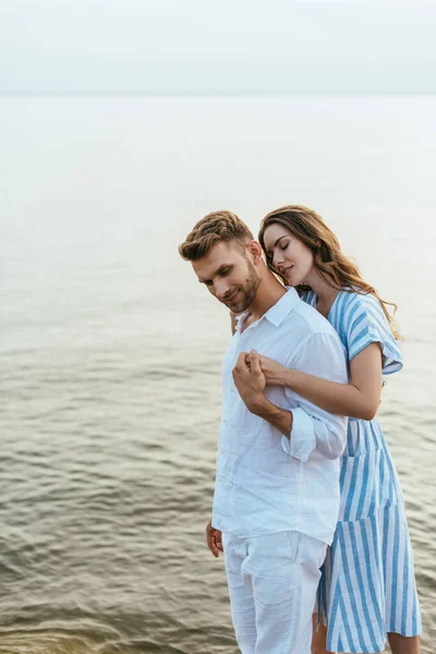 Atractiva novia abrazando y cogido de la mano con guapo novio cerca de river - foto de stock