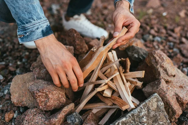 Vue recadrée de jeune homme touchant bâtons tout en faisant feu de joie près de pierres — Photo de stock