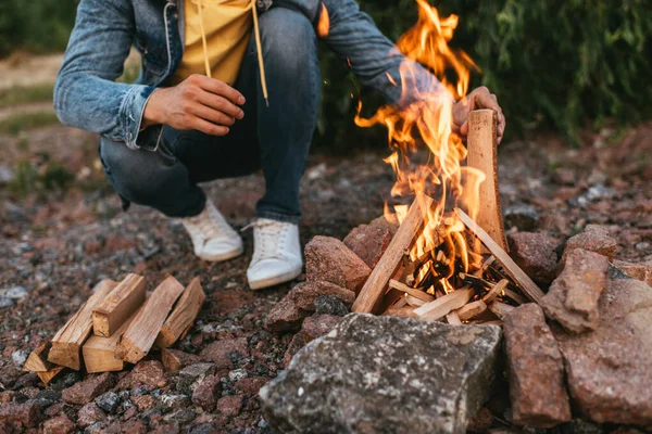 Cropped view of man sitting and putting log in burning bonfire — Stock Photo