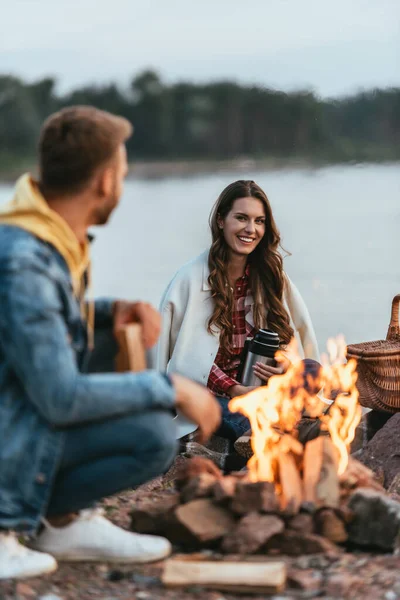 Selective focus of happy woman holding thermos and looking at boyfriend near bonfire — Stock Photo