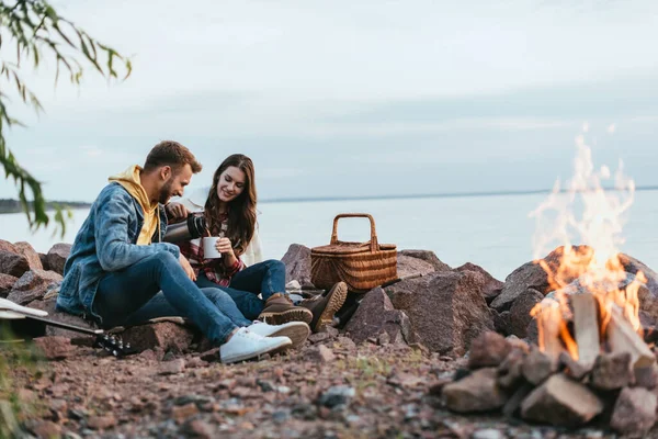 Selective focus of happy woman pouring tea in cup near cheerful man and burning bonfire — Stock Photo