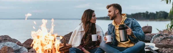 Panoramic shot of couple holding cups and sitting near bonfire and lake — Stock Photo