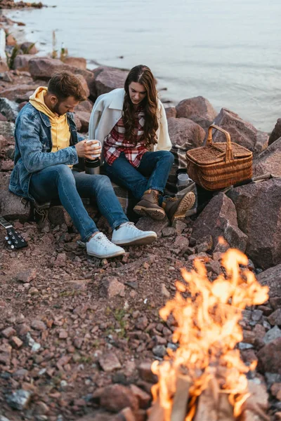 Selective focus of couple sitting on stones near bonfire and lake — Stock Photo