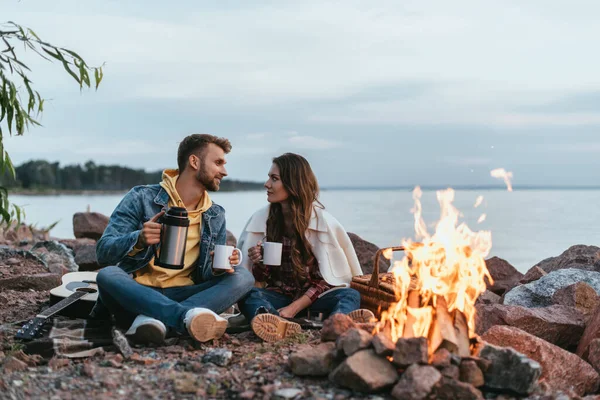 Selektiver Fokus des Paares, das Tassen hält und auf Felsen in der Nähe von Lagerfeuer und See sitzt — Stockfoto