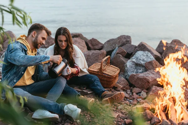 Selektiver Fokus eines gutaussehenden Mannes, der Tee in eine Tasse in der Nähe von Mädchen, See und Lagerfeuer gießt — Stockfoto