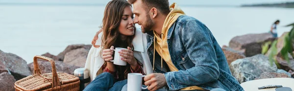 Panoramic concept of happy couple holding cups near river — Stock Photo