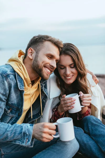 Feliz hombre abrazando alegre joven mujer y sosteniendo taza de té - foto de stock