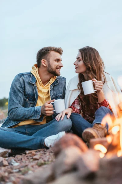 Enfoque selectivo de hombre feliz y mujer joven sosteniendo tazas de té cerca de la hoguera - foto de stock