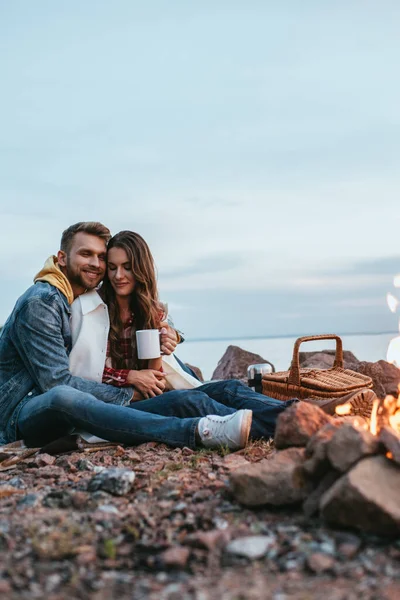 Selective focus of happy man hugging girl with cup near bonfire — Stock Photo
