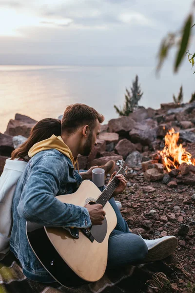 Foco seletivo do homem barbudo tocando guitarra acústica perto de namorada, rio e fogueira — Fotografia de Stock