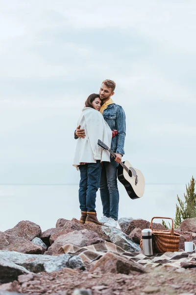 Guapo hombre sosteniendo la guitarra acústica y abrazando a la chica mientras está de pie cerca del río - foto de stock
