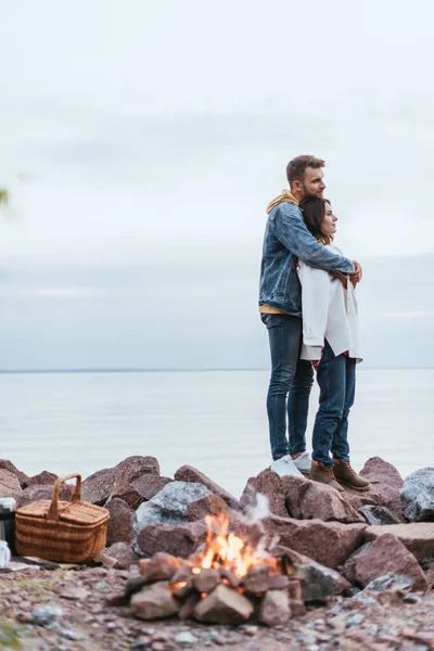 Selektiver Fokus eines gutaussehenden Mannes, der attraktive Frau umarmt, während er in der Nähe von Fluss und Lagerfeuer steht — Stockfoto