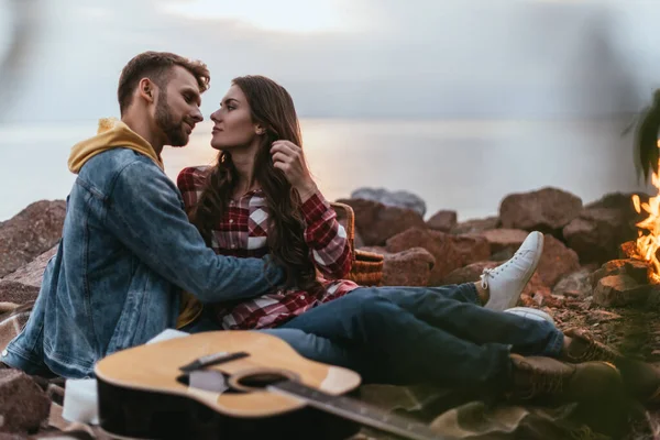 Селективное внимание бородатого мужчины, обнимающего подругу возле акустической гитары и костра — стоковое фото