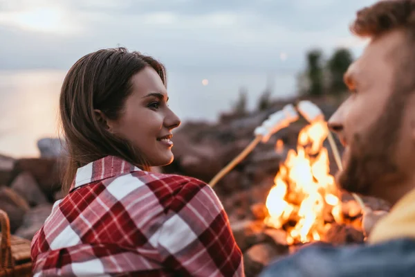 Foco seletivo de mulher feliz olhando para namorado perto de fogueira e marshmallows em paus — Fotografia de Stock