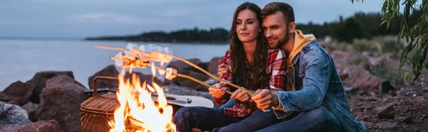 Panoramic shot of couple roasting marshmallows on sticks near bonfire — Stock Photo