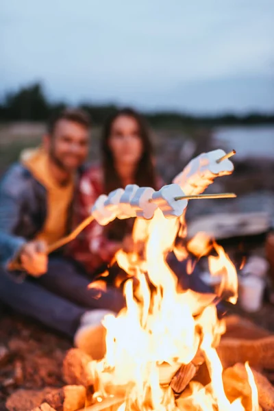 Selective focus of puffy marshmallows on sticks near burning bonfire and couple — Stock Photo