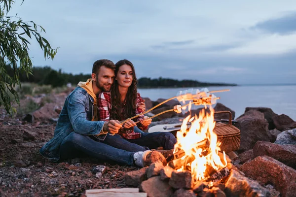 Feliz pareja asando malvaviscos en palos cerca de la hoguera - foto de stock